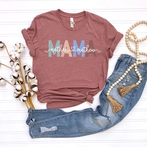 May include: A maroon t-shirt with the word "MAMA" in pastel colors and the names "nathan" and "matthew" in white script. The shirt is paired with blue jeans and a beaded necklace with tassels. Cotton stems are also in the image.