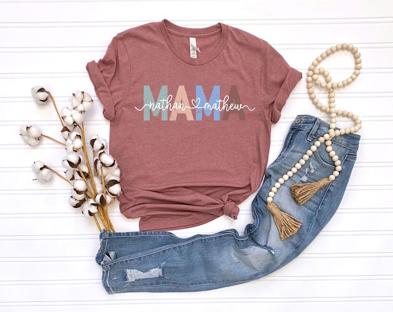 May include: A maroon t-shirt with the word "MAMA" in pastel colors and the names "nathan" and "matthew" in white script. The shirt is paired with blue jeans and a beaded necklace with tassels. Cotton stems are also in the image.