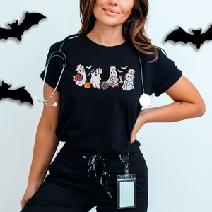May include: A black t-shirt featuring a graphic of four ghosts dressed as nurses. The ghosts are holding medical supplies and a pumpkin. A stethoscope is draped over the shoulder.