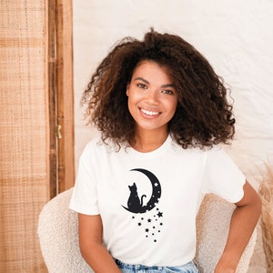 May include: A white t-shirt with a black silhouette of a cat sitting on a crescent moon with stars. The text "Ojev Apparel Soft Quality Clothing" is in the top right corner of the image.