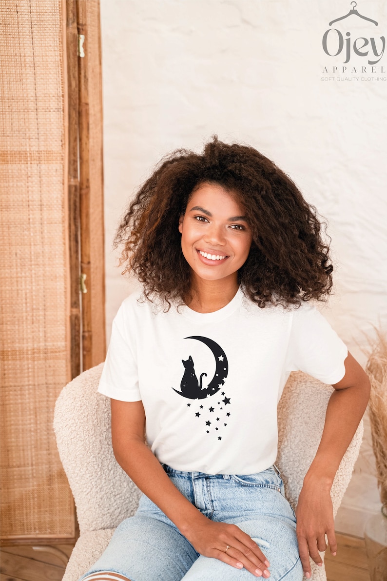 May include: A white t-shirt with a black silhouette of a cat sitting on a crescent moon with stars. The text "Ojev Apparel Soft Quality Clothing" is in the top right corner of the image.
