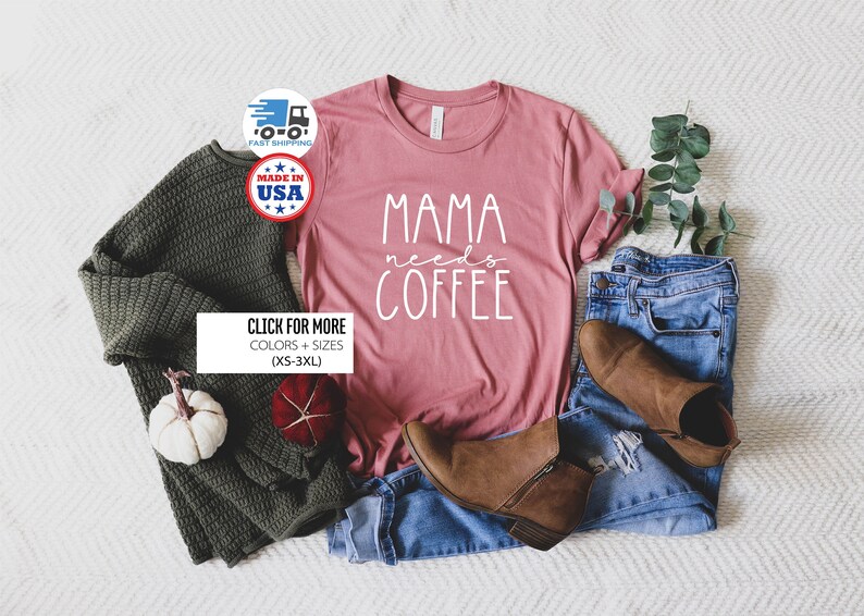May include: A pink t-shirt with the text "Mama needs coffee" in white lettering. The shirt is laid out on a white surface with a pair of jeans, a sweater, and a small white pumpkin.