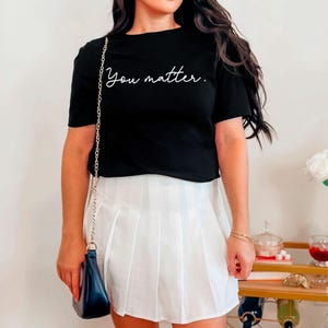 May include: A black cropped t-shirt with the text "You matter." in white script. The shirt is paired with a white pleated skirt.