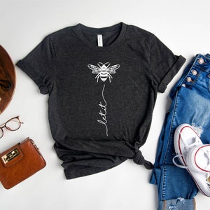 May include: A dark gray t-shirt with a white bee graphic and the text "let it bee" printed on the front. The shirt is laid flat on a white surface with a pair of jeans, a brown purse, a pair of glasses, and a camera.