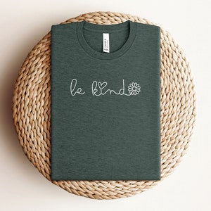 Puede incluir: Una camiseta verde oscuro con el texto "be kind" en blanco con un peque&ntilde;o coraz&oacute;n y un gr&aacute;fico de flor.