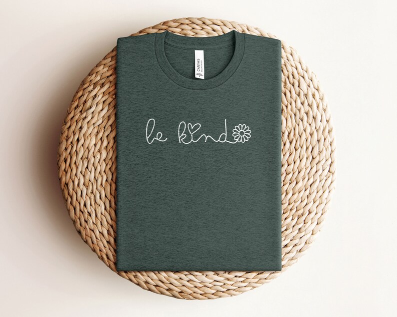 Puede incluir: Una camiseta verde oscuro con el texto "be kind" en blanco con un peque&ntilde;o coraz&oacute;n y un gr&aacute;fico de flor.