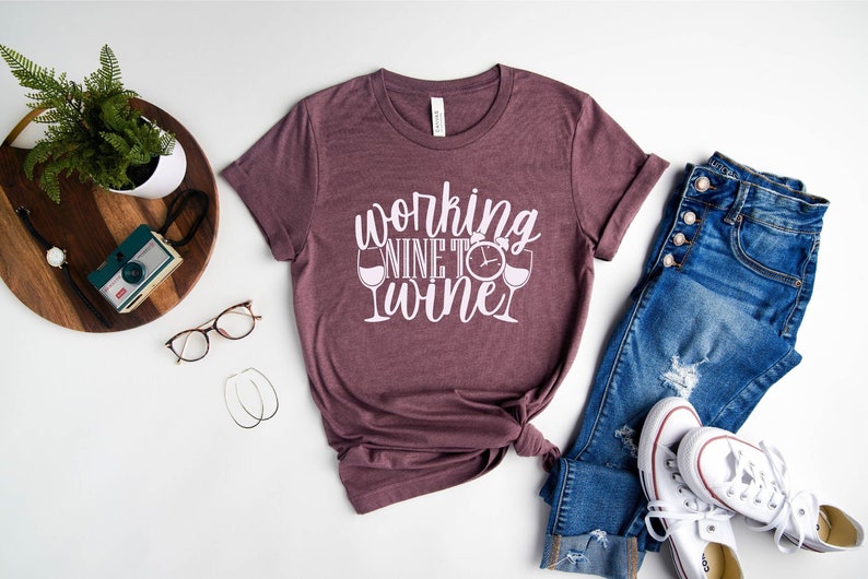 May include: A maroon t-shirt with the text "working nine to wine" in white lettering. The shirt features a graphic of two wine glasses with a clock face in between.