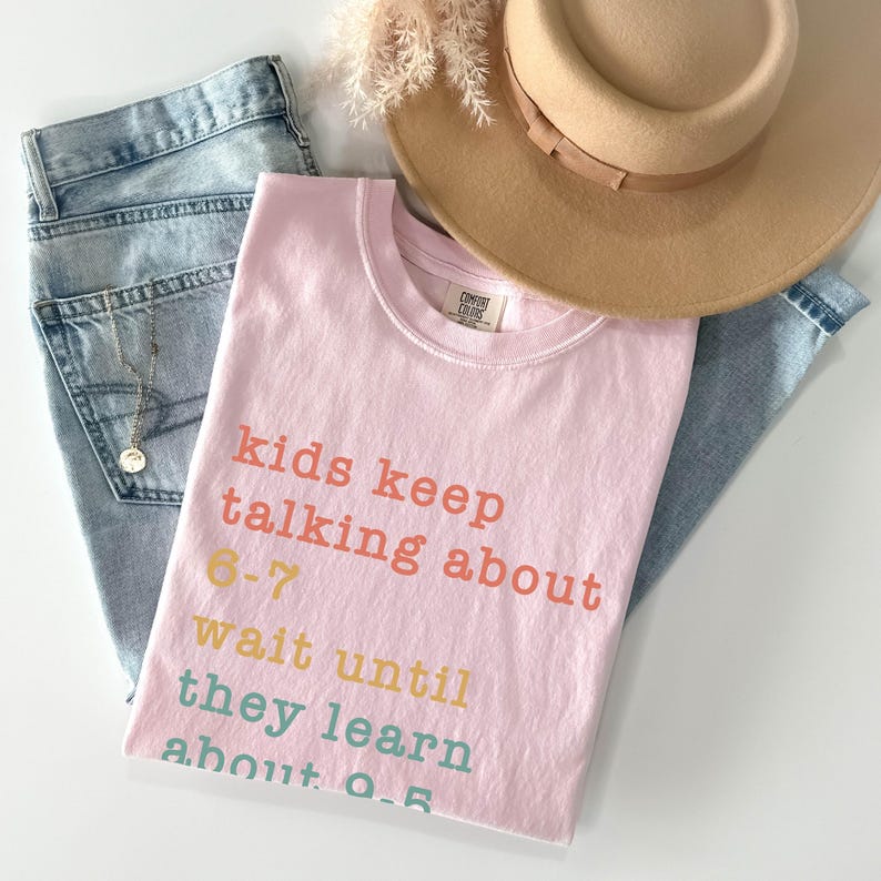 May include: A pale pink t-shirt with the text "kids keep talking about 6-7 wait until they learn about 0-5" in various colours. The shirt is laid on top of light blue jeans, a tan hat, and a necklace.