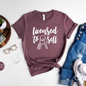 Pode incluir: Camiseta de cor marrom heather com o texto "Licensed to Sell" e um par de chaves com a hashtag "#realtor" impressa em branco.