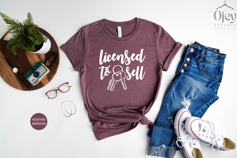 Pode incluir: Camiseta de cor marrom heather com o texto "Licensed to Sell" e um par de chaves com a hashtag "#realtor" impressa em branco.