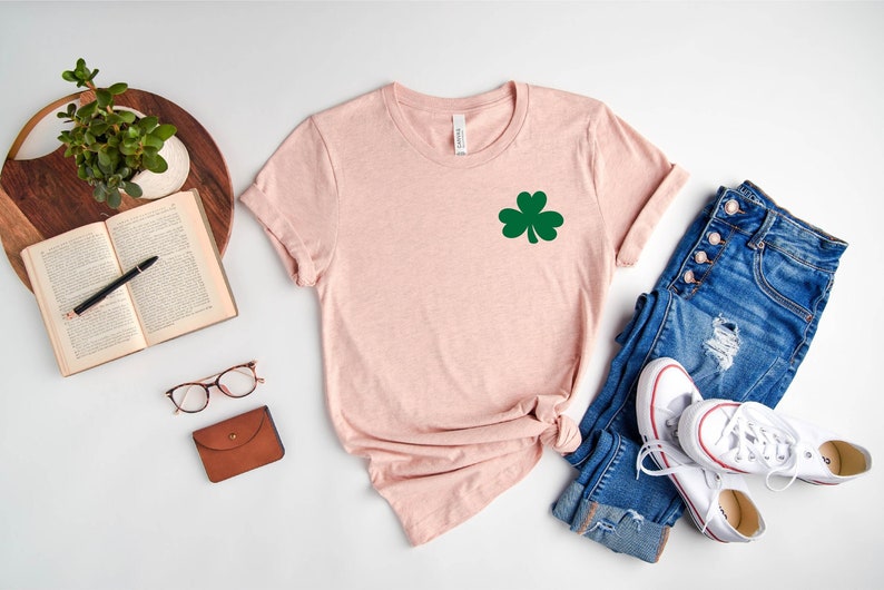 May include: A pink t-shirt with a green shamrock design. The shirt is laid out on a white surface with a pair of jeans, a book, a pair of glasses, and a small brown wallet.