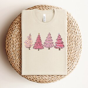 May include: A beige t-shirt featuring a design of four pink Christmas trees with white lights and gold stars on a beige background.