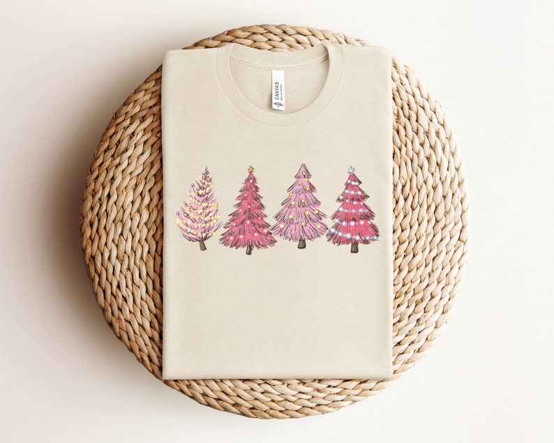May include: A beige t-shirt featuring a design of four pink Christmas trees with white lights and gold stars on a beige background.