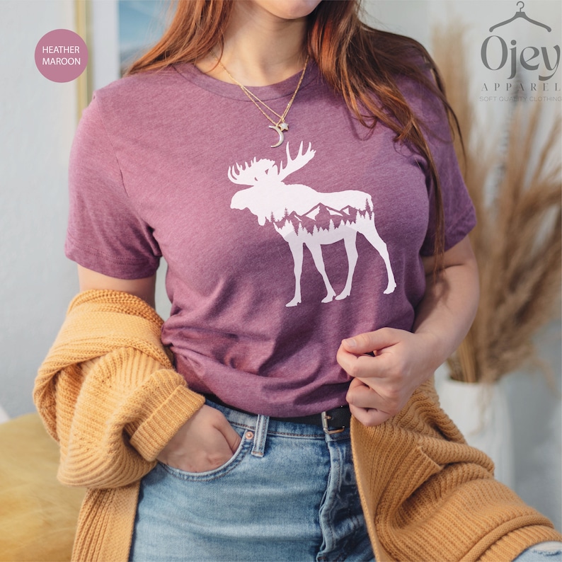 May include: Heather maroon t-shirt with a white moose and mountain graphic. The moose silhouette has a mountain and tree design inside. The shirt is short-sleeved and made of a soft material. The text "OjeY Apparel" is in the upper right corner.