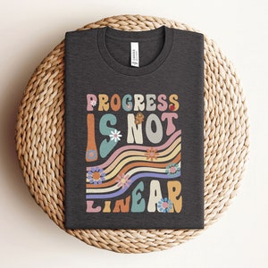 May include: A dark gray t-shirt with a colorful graphic that says "Progress is not linear" in a retro font. The graphic features wavy lines and flowers.