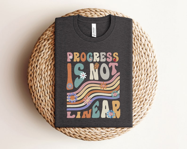 May include: A dark gray t-shirt with a colorful graphic that says "Progress is not linear" in a retro font. The graphic features wavy lines and flowers.