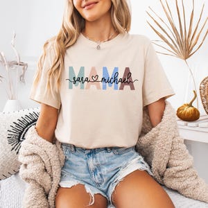 May include: Beige t-shirt with the word "MAMA" in colorful block letters and the names "Sara" and "Michael" below. The shirt has a crew neck and short sleeves. The text is in a variety of colors, including blue, pink, and brown.