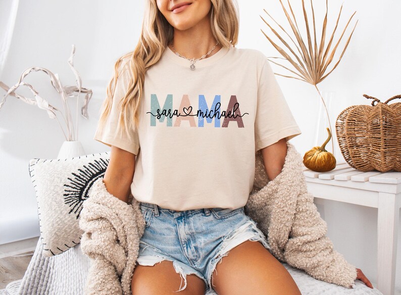 May include: Beige t-shirt with the word "MAMA" in colorful block letters and the names "Sara" and "Michael" below. The shirt has a crew neck and short sleeves. The text is in a variety of colors, including blue, pink, and brown.