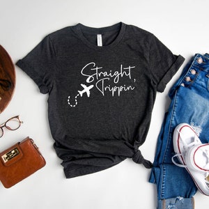 May include: Heather dark gray t-shirt with the text "Straight Trippin'" and a white airplane graphic. The shirt is styled with blue jeans, white sneakers, and a brown purse.