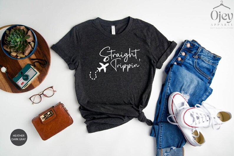 May include: Heather dark gray t-shirt with the text "Straight Trippin'" and a white airplane graphic. The shirt is styled with blue jeans, white sneakers, and a brown purse.