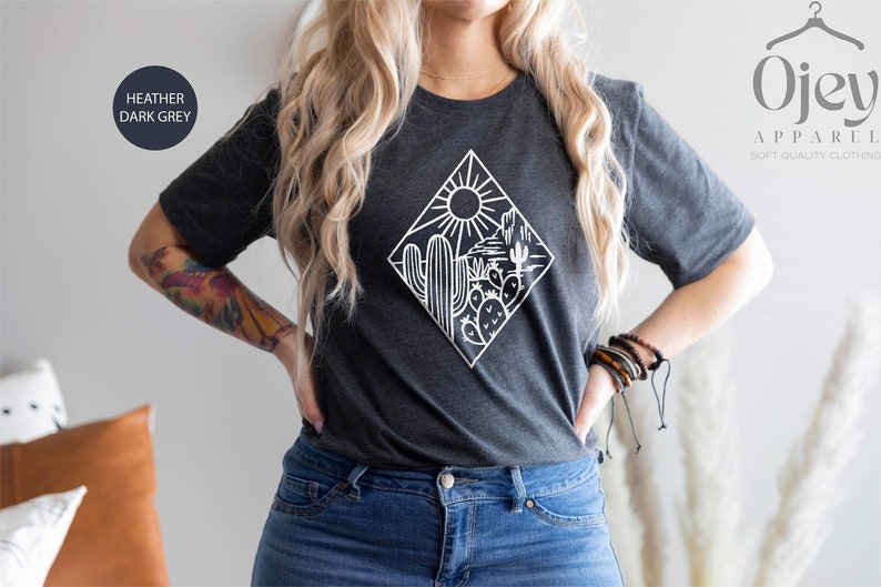 May include: A heather dark grey t-shirt with a white graphic of a desert scene with a sun, cactus, and mountains. The text "Ojev Apparel Soft Quality Clothing" is in the top right corner of the image.