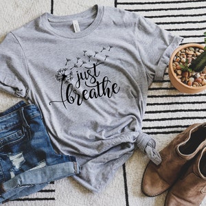 K&ouml;nnte beinhalten: Ein graues T-Shirt mit einer L&ouml;wenzahndarstellung und dem Text "just breathe" in schwarzer Schrift.