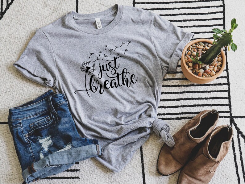 K&ouml;nnte beinhalten: Ein graues T-Shirt mit einer L&ouml;wenzahndarstellung und dem Text "just breathe" in schwarzer Schrift.