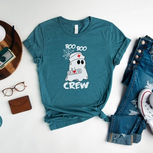May include: A teal blue t-shirt featuring a white ghost wearing a nurse's hat and holding a bandage. The text "Boo Boo Crew" is printed on the shirt.