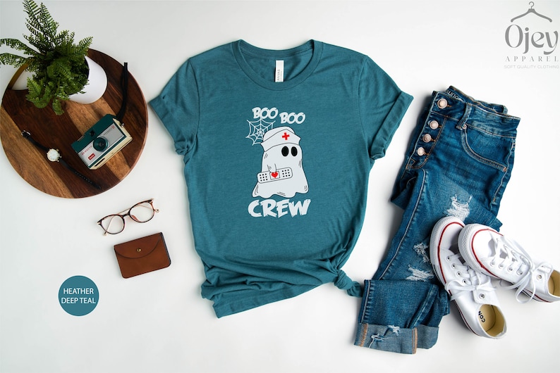 May include: A teal blue t-shirt featuring a white ghost wearing a nurse's hat and holding a bandage. The text "Boo Boo Crew" is printed on the shirt.