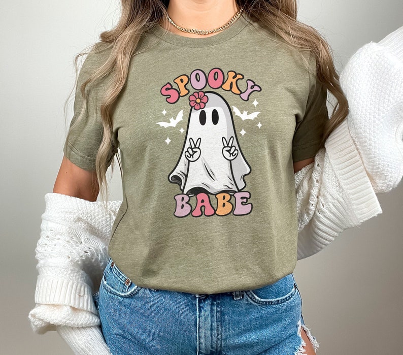 May include: A green t-shirt with a cartoon ghost wearing a flower on its head. The ghost has peace signs for hands and the text "Spooky Babe" in rainbow letters.