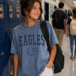 May include: A blue short-sleeved crew neck t-shirt featuring the word EAGLES in dark blue block lettering above a graphic of an eagle head. The shirt has a faded, vintage-style wash and is worn with a black backpack over one shoulder.