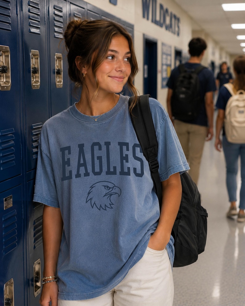 May include: A blue short-sleeved crew neck t-shirt featuring the word EAGLES in dark blue block lettering above a graphic of an eagle head. The shirt has a faded, vintage-style wash and is worn with a black backpack over one shoulder.