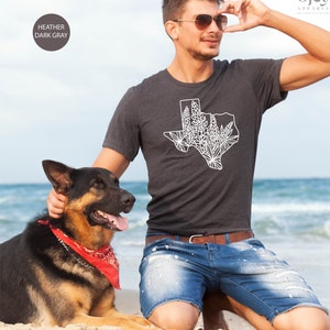May include: Heather dark gray t-shirt featuring a white graphic of the state of Texas with a floral design. The shirt is worn by a person on a beach with a dog. The shirt is from Ojev Apparel.