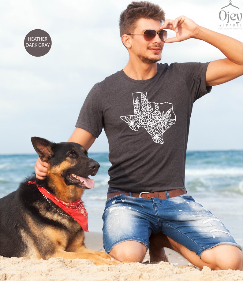 May include: Heather dark gray t-shirt featuring a white graphic of the state of Texas with a floral design. The shirt is worn by a person on a beach with a dog. The shirt is from Ojev Apparel.
