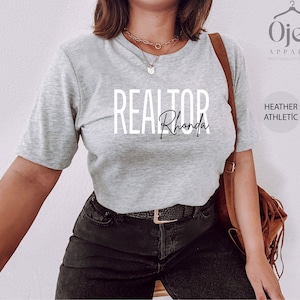 Peut inclure: Un t-shirt gris chin&eacute; avec le texte "REALTOR Rhonda" imprim&eacute; sur le devant. Le t-shirt est port&eacute; par une personne avec un jean noir et un sac en cuir marron.