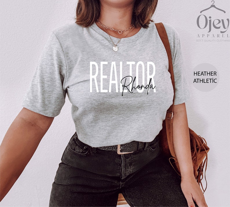 Peut inclure: Un t-shirt gris chin&eacute; avec le texte "REALTOR Rhonda" imprim&eacute; sur le devant. Le t-shirt est port&eacute; par une personne avec un jean noir et un sac en cuir marron.