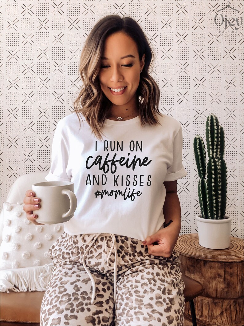 K&ouml;nnte beinhalten: Wei&szlig;es T-Shirt mit schwarzem Text, der "I run on caffeine and kisses #momlife" lautet. Eine Person tr&auml;gt das Shirt und h&auml;lt eine wei&szlig;e Tasse.