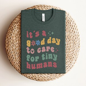 May include: A dark green t-shirt with the text "it's a good day to care for tiny humans" in colorful, retro-style lettering.
