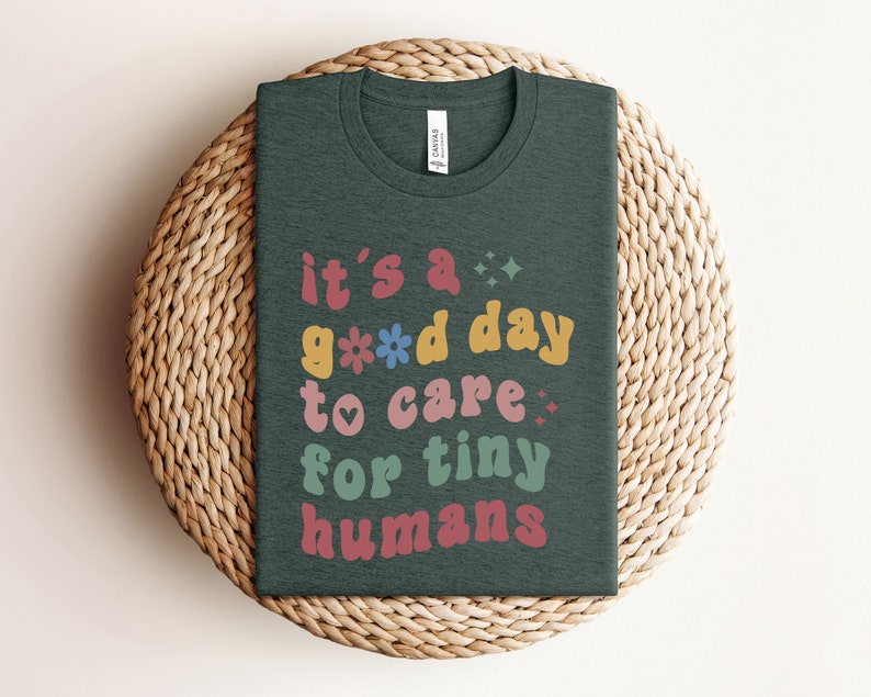May include: A dark green t-shirt with the text "it's a good day to care for tiny humans" in colorful, retro-style lettering.