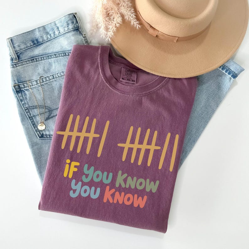 May include: A mauve-colored t-shirt with the text "If You Know You Know" in colorful letters. The shirt is folded on top of a pair of light blue jeans, a beige hat, and a gold necklace.