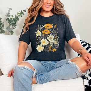 K&ouml;nnte beinhalten: Eine Frau tr&auml;gt ein dunkelgraues T-Shirt mit einem Blumendruck aus gelben und wei&szlig;en Blumen. Das Shirt hat eine lockere Passform und eine verk&uuml;rzte L&auml;nge. Die Frau sitzt auf einem wei&szlig;en Sofa mit einer schwarz-wei&szlig;en karierten Wolldecke.