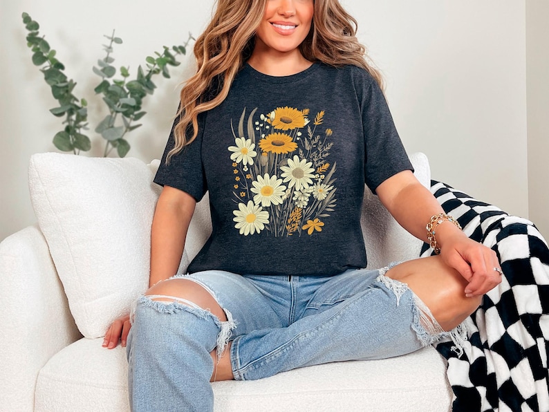K&ouml;nnte beinhalten: Eine Frau tr&auml;gt ein dunkelgraues T-Shirt mit einem Blumendruck aus gelben und wei&szlig;en Blumen. Das Shirt hat eine lockere Passform und eine verk&uuml;rzte L&auml;nge. Die Frau sitzt auf einem wei&szlig;en Sofa mit einer schwarz-wei&szlig;en karierten Wolldecke.