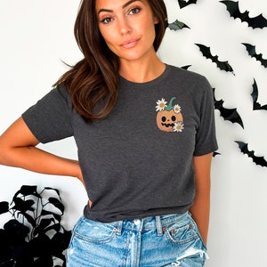 May include: A dark gray t-shirt with a cute pumpkin design. The pumpkin has a friendly face and is decorated with white flowers.