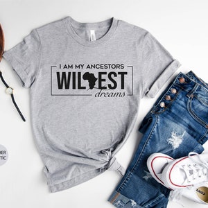 Peut inclure: T-shirt gris avec le texte "I am my ancestors wildest dreams" et un contour noir du continent africain. Le t-shirt est pos&eacute; sur une surface blanche avec un jean, une paire de baskets blanches et une paire de lunettes.