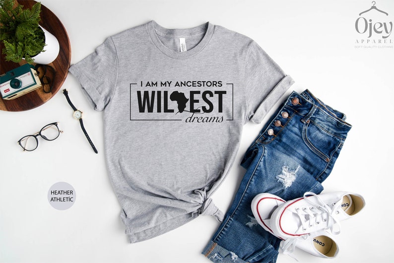Peut inclure: T-shirt gris avec le texte "I am my ancestors wildest dreams" et un contour noir du continent africain. Le t-shirt est pos&eacute; sur une surface blanche avec un jean, une paire de baskets blanches et une paire de lunettes.