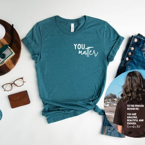 May include: A teal heather t-shirt with the text "YOU matter" in white. The shirt is shown on a white background with a pair of glasses, a brown wallet, and a wooden tray with a camera and a watch. A second image shows a black t-shirt with the text "TO THE PERSON BEHIND ME: YOU ARE AMAZING. BEAUTIFUL AND ENOUGH. Remember that" in white. The t-shirt is being worn by a person with long brown hair standing on a beach.