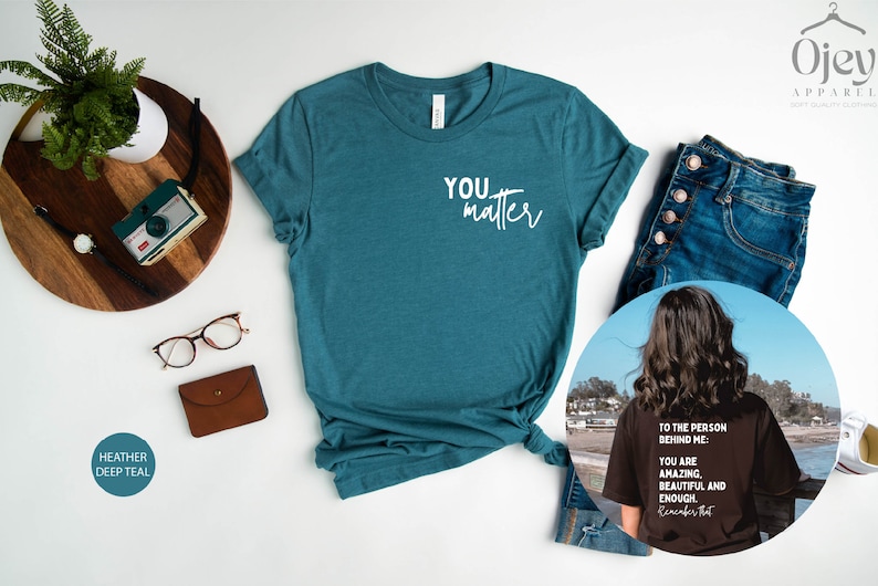 May include: A teal heather t-shirt with the text "YOU matter" in white. The shirt is shown on a white background with a pair of glasses, a brown wallet, and a wooden tray with a camera and a watch. A second image shows a black t-shirt with the text "TO THE PERSON BEHIND ME: YOU ARE AMAZING. BEAUTIFUL AND ENOUGH. Remember that" in white. The t-shirt is being worn by a person with long brown hair standing on a beach.