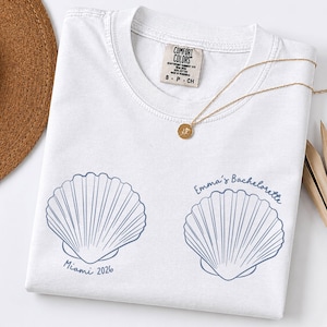 May include: A white t-shirt featuring two blue line-art seashell illustrations. The left shell is printed with the text "Miami 2026" and the right shell is printed with the text "Emma's Bachelorette". The shirt has a Comfort Colors brand neck label.