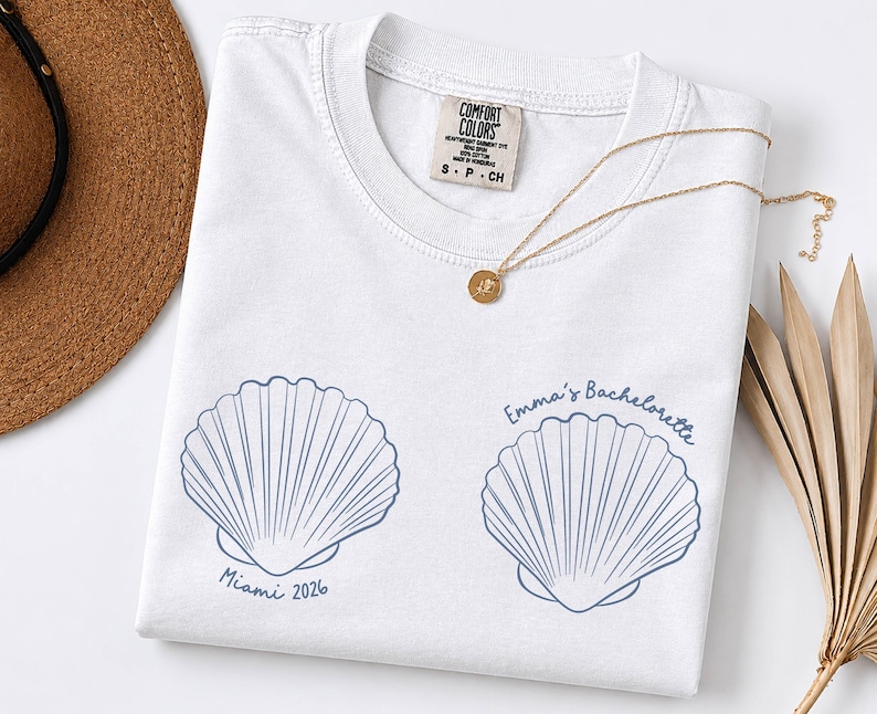May include: A white t-shirt featuring two blue line-art seashell illustrations. The left shell is printed with the text "Miami 2026" and the right shell is printed with the text "Emma's Bachelorette". The shirt has a Comfort Colors brand neck label.