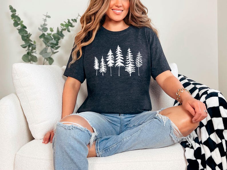 May include: A woman wearing a dark gray t-shirt with a white silhouette of a forest on the front. She is sitting on a white couch with a black and white checkered throw blanket.